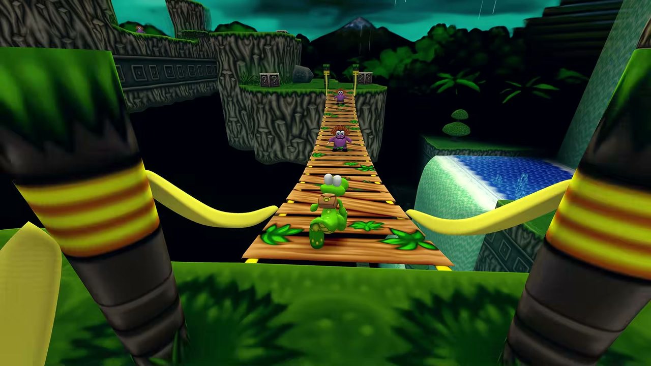 Croc and two Gobbos stand on a rope bridge over water, with mountains and a dark sky behind